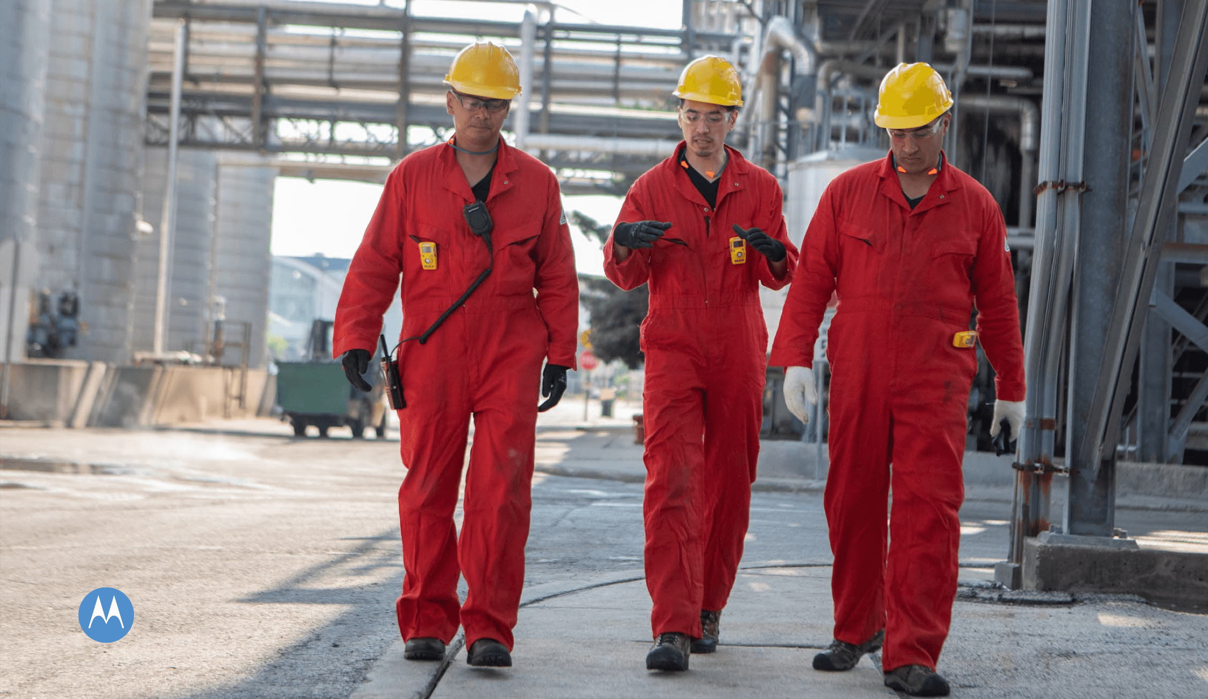 Tres trabajadores con overoles rojos y cascos amarillos caminan por una instalación industrial exterior.