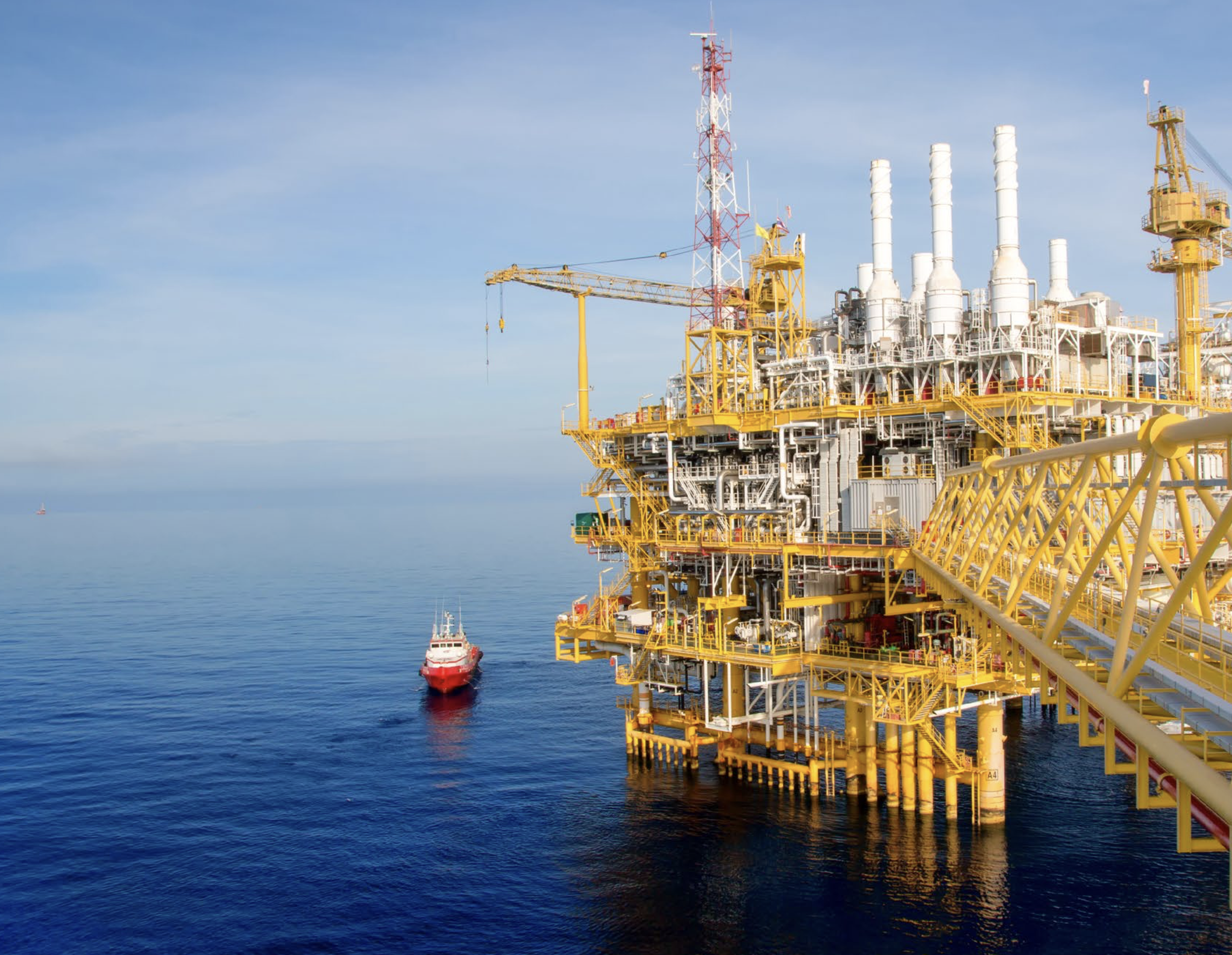 Plataforma petrolífera amarilla con chimeneas blancas y un barco rojo en el mar azul.